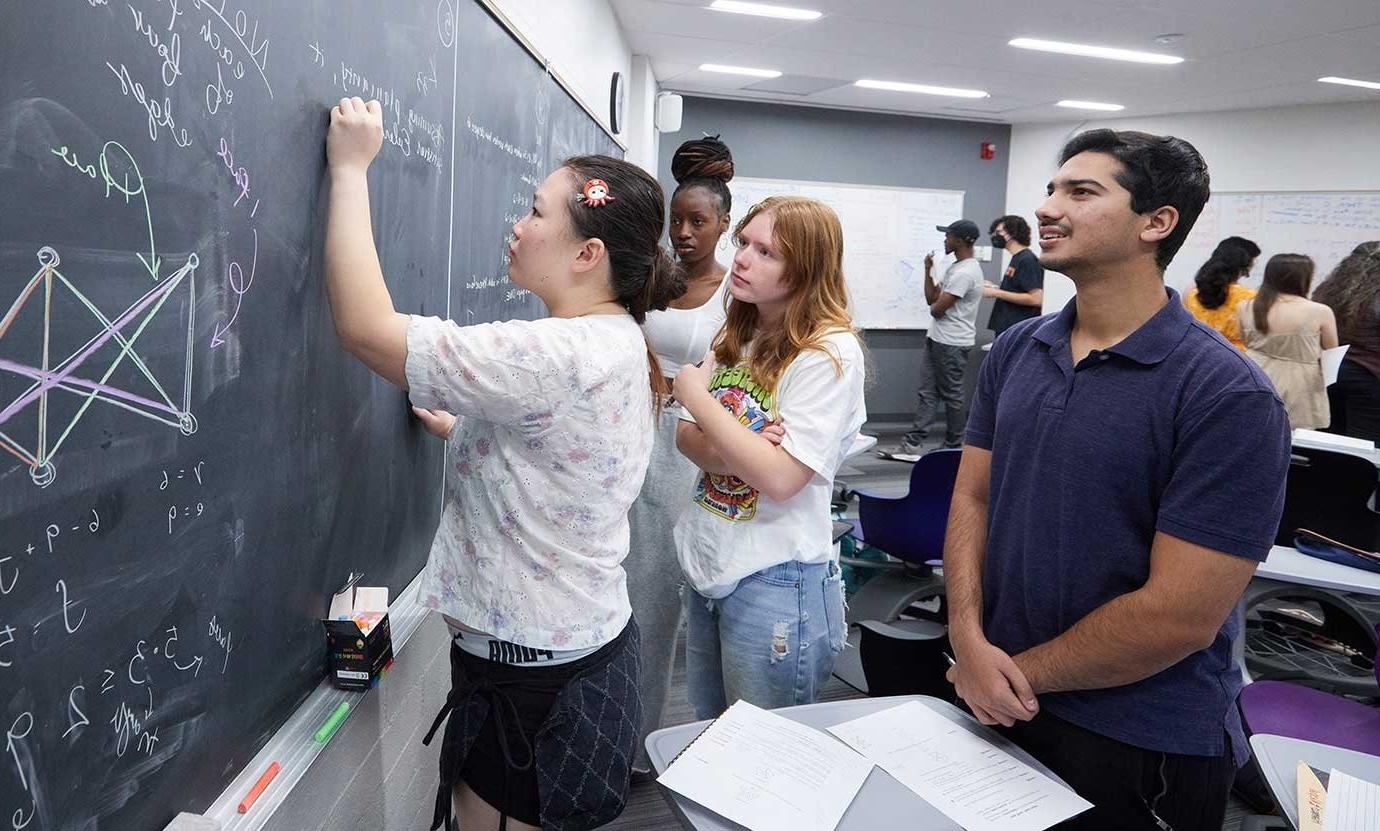 Student writing with chalk on blackboard while others watch